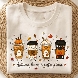 May include: A cream-coloured t-shirt featuring a graphic of four cartoon coffee cups with faces, ghosts, and pumpkins. The text "Hello Fall" is on one cup, and the phrase "Autumn leaves & coffee please" is printed in black script at the bottom.