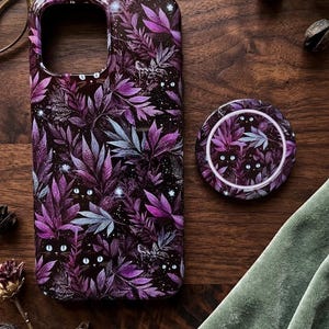 May include: A rectangular smartphone case and a circular phone grip featuring a dark pattern of purple and black foliage with hidden black cat faces and bright white eyes peering out from the leaves, set against a deep black background with small white stars.