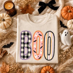 May include: A cream-coloured crewneck sweatshirt featuring the word BOO in large, decorative letters. The B is black and white buffalo plaid, the O is pink with white ghosts and black bats, and the second O has vertical orange, purple, and white stripes.