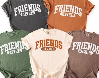 Könnte beinhalten: Fünf kurzärmelige T-Shirts mit Rundhalsausschnitt in Anthrazit, Orange, Olivgrün, Creme und Braun. Jedes Shirt zeigt den Schriftzug FRIENDS GIVING in einer abgenutzten, gebogenen College-Schrift auf der Brust, wobei das Wort GIVING in einem Rechteck steht.
