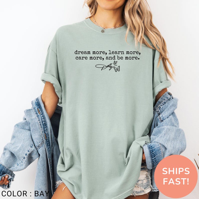 May include: A sage green short-sleeve t-shirt featuring the black printed text "dream more, learn more, care more, and be more." with a signature and two small butterfly illustrations. The shirt is worn with a light blue denim jacket over the shoulders.