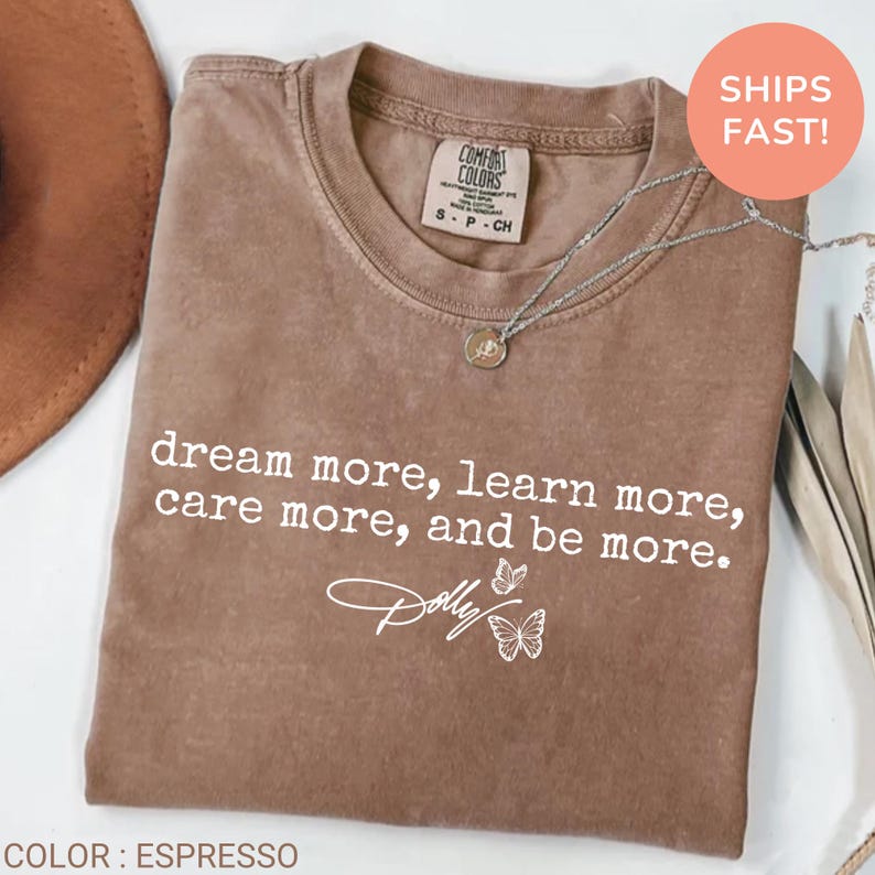 May include: An espresso-colored short-sleeve t-shirt featuring the white printed text "dream more, learn more, care more, and be more." along with a script "Dolly" logo and two butterfly outlines. The shirt has a crew neckline and a size S label.
