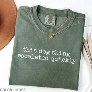Puede incluir: Una camiseta de manga corta color verde musgo con cuello redondo y el texto blanco "this dog thing escalated quickly" impreso en el frente. La camiseta tiene una etiqueta de la marca Comfort Colors en el cuello interior con la talla S, P, CH.