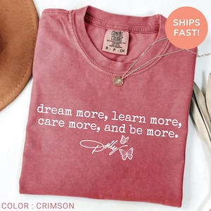 May include: A crimson short-sleeved t-shirt featuring the white printed text "dream more, learn more, care more, and be more." and a signature graphic with two butterflies. The shirt has a crew neckline and a Comfort Colors brand label at the neck.