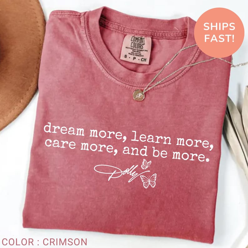 May include: A crimson short-sleeved t-shirt featuring the white printed text "dream more, learn more, care more, and be more." and a signature graphic with two butterflies. The shirt has a crew neckline and a Comfort Colors brand label at the neck.