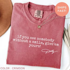 May include: A crimson short-sleeved t-shirt featuring the white printed text: if you see somebody without a smile, give'em yours! and the signature Dolly. The shirt has a crew neckline and a Comfort Colors brand label visible at the inner collar.
