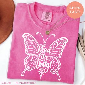 May include: A pink short-sleeve t-shirt featuring a large white graphic of a butterfly with intricate line work. The text Read Like Dolly! is printed in white in the center of the butterfly. The shirt has a crew neckline and a tag.