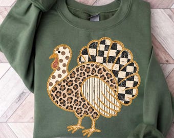 Könnte beinhalten: Ein militärgrünes Sweatshirt mit Rundhalsausschnitt und einer großen Truthahn-Grafik. Der Truthahn hat einen Hals mit Punkten, einen Körper mit Leopardenmuster, einen gestreiften Flügel und einen schwarz-weiß karierten Schwanz, alles mit goldenem Glitzer umrandet.
