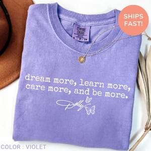May include: A violet short-sleeved t-shirt with a crew neckline. The shirt features white text that reads: dream more, learn more, care more, and be more. Below the text is a white script signature that reads Dolly and two small butterfly outlines.