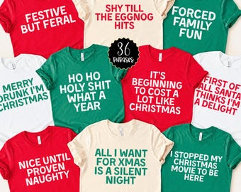 Könnte beinhalten: Eine Sammlung von kurzärmeligen T-Shirts in Rot, Grün und Creme, die jeweils mit einzigartigen weihnachtlichen Texten wie Festive But Feral, Shy Till The Eggnog Hits, Forced Family Fun und anderen humorvollen Weihnachtssprüchen bedruckt sind.