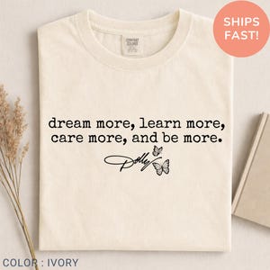 May include: An ivory t-shirt featuring the black text "dream more, learn more, care more, and be more." below which is a script signature reading "Dolly" accompanied by two small black butterfly illustrations centered on the chest area.
