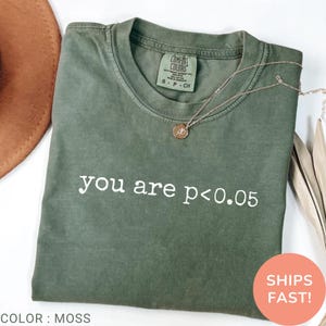 Op de afbeelding: Een mosgroen t-shirt met korte mouwen en de witte opdruk you are p<0.05 op de borst. Het shirt heeft een ronde hals met een Comfort Colors merklabel aan de binnenkant en een dunne goudkleurige ketting die bovenop het shirt ligt.
