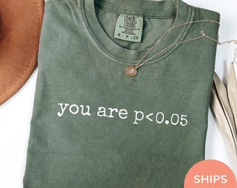 Könnte beinhalten: Ein moosgrünes Kurzarm-T-Shirt mit dem weißen Aufdruck you are p<0.05 auf der Brust. Das Shirt hat einen Rundhalsausschnitt mit einem Comfort Colors Markenetikett im Kragen und eine dünne goldfarbene Halskette, die darauf platziert ist.