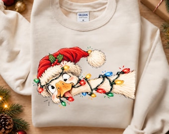 Könnte beinhalten: Ein sandfarbenes Sweatshirt mit Rundhalsausschnitt und einer Grafik einer Gans, die eine rote Weihnachtsmütze mit weißem Bommel und Stechpalmenblättern trägt. Die Gans ist in eine Lichterkette verheddert. Das Innenetikett zeigt Gildan M.