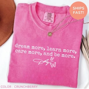 May include: A bright pink short-sleeved t-shirt with a crew neckline. The shirt features white text that reads, dream more, learn more, care more, and be more, followed by a signature that says Dolly and two small white butterfly illustrations.