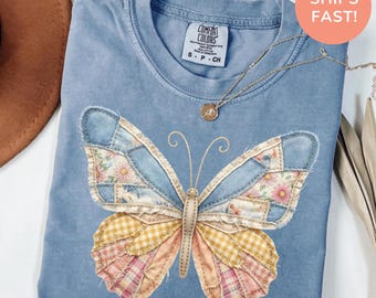 Könnte beinhalten: Ein blaues Comfort Colors T-Shirt mit einem Patchwork-Schmetterlingsdesign. Die Schmetterlingsflügel bestehen aus Jeans-, Blumen- und Karostoffen. Eine goldene Halskette mit Anhänger liegt auf dem Shirt. Das Shirt ist mit "S - P - CH" gekennzeichnet.