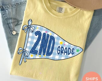 Könnte beinhalten: Ein blassgelbes T-Shirt mit einer Grafik eines hellblau-weiß karierten Wimpels. Der Wimpel zeigt den dunkelblauen Schriftzug 2ND GRADE und ist mit kleinen gelben und grünen Sternen verziert. Der Wimpel ist mit zwei Schleifen an einem Stab befestigt.