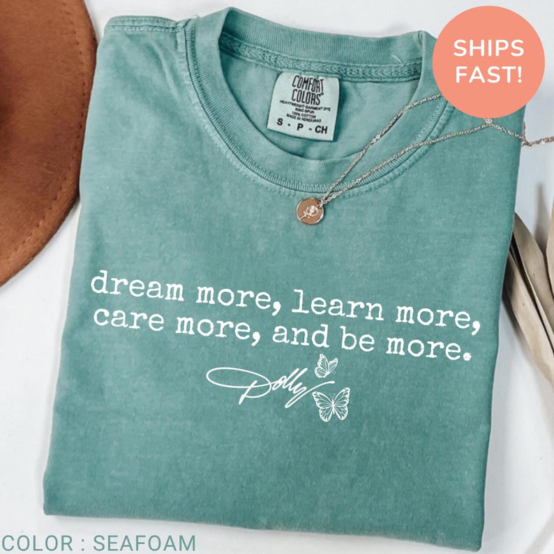 May include: A seafoam green short-sleeve t-shirt featuring the white printed text "dream more, learn more, care more, and be more." with a signature style "Dolly" logo and two butterfly illustrations below the text, plus a silver pendant necklace.
