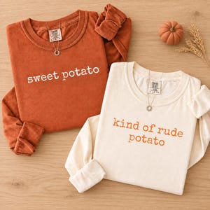 Könnte beinhalten: Zwei langärmelige Rundhals-Shirts. Eines ist rostorange mit weißem Text 'sweet potato'. Das andere ist cremefarben mit orangefarbenem Text 'kind of rude potato'. Beide Shirts haben ein Comfort Colors-Etikett am Halsausschnitt.