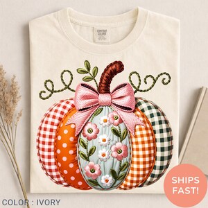 May include: An ivory t-shirt featuring a large graphic of a pumpkin with a pink bow. The pumpkin has segments with red gingham, orange polka dots, a central light blue section with pink and white flowers, and green gingham patterns.