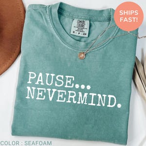 May include: A seafoam green short-sleeve t-shirt featuring the white text PAUSE... NEVERMIND. printed on the front. The shirt has a crew neckline and a Comfort Colors brand label inside the collar. A silver-toned necklace rests on the fabric.