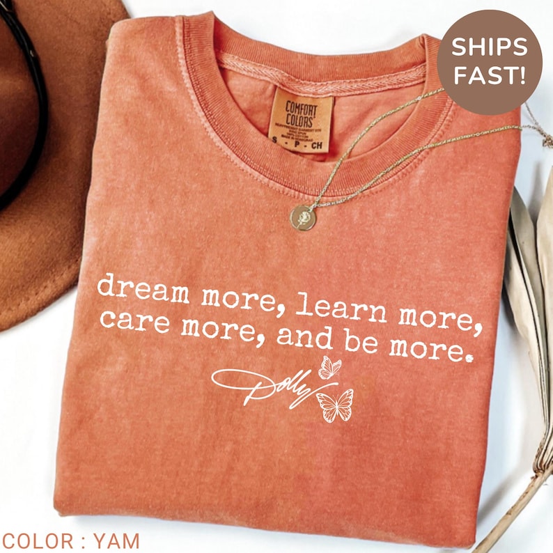 May include: A yam-colored short-sleeve t-shirt featuring the white text "dream more, learn more, care more, and be more." and a signature graphic with two butterflies. The shirt has a Comfort Colors brand label on the inner neckline.