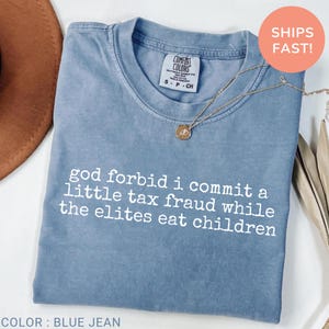 May include: A blue jean colored t-shirt with a crew neckline and short sleeves. The shirt features white text printed on the front that reads: god forbid i commit a little tax fraud while the elites eat children. The brand label reads Comfort Colors.