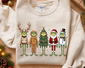 Könnte beinhalten: Ein sandfarbenes Sweatshirt mit Rundhalsausschnitt und einem Aufdruck von fünf grünen Fröschen in Weihnachtskostümen: ein Rentier, ein Lebkuchenmann, ein Weihnachtsbaum, der Weihnachtsmann und ein Schneemann. Das Gildan-Etikett ist am Hals sichtbar.