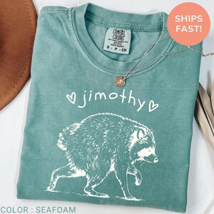 May include: A seafoam green short-sleeve t-shirt featuring a white graphic of a raccoon walking. Above the raccoon, the text reads jimothy with a small heart on each side. The shirt has a crew neckline and a Comfort Colors brand label inside.