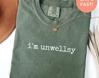 Könnte beinhalten: Ein moosgrünes T-Shirt mit Rundhalsausschnitt und dem weißen Schriftzug i'm unwellsy auf der Brust. Das Shirt hat ein Comfort Colors Markenetikett am inneren Halsausschnitt und wird mit einer dünnen goldfarbenen Halskette gezeigt.