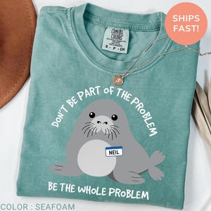 May include: A seafoam green t-shirt featuring a graphic of a gray seal with a white name tag that reads NEIL. The text above the seal reads DON'T BE PART OF THE PROBLEM and the text below the seal reads BE THE WHOLE PROBLEM in white lettering.