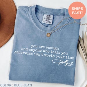 May include: A blue jean colored t-shirt with a crew neckline and short sleeves. The shirt features white text that reads, you are enough and anyone who tells you otherwise isn't worth your time, followed by a signature that reads Dolly xoxo.