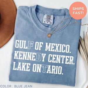 Op de afbeelding: Een kort mouwloos t-shirt in de kleur spijkerblauw met witte tekst die luidt GULF OF MEXICO. KENNEDY CENTER. LAKE ONTARIO. Het shirt heeft een ronde hals en een Comfort Colors merklabel aan de binnenkant. Een gouden ketting ligt op de stof.