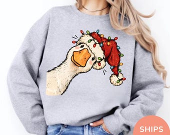 Könnte beinhalten: Ein hellgraues Sweatshirt mit Rundhalsausschnitt und einem Grafikdruck einer weißen Gans, die eine rote Weihnachtsmütze mit bunten Lichterketten trägt. Die Gans hat einen orangefarbenen Schnabel und rosa Wangen auf der Vorderseite.