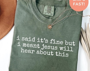 Könnte beinhalten: Ein moosgrünes T-Shirt mit Rundhalsausschnitt und dem weißen Text "i said it's fine but i meant jesus will hear about this" auf der Vorderseite. Das Shirt hat ein Comfort Colors Markenetikett am inneren Halsausschnitt und eine dünne Goldkette.