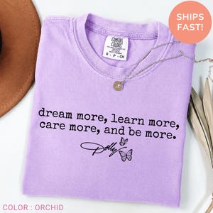 May include: A light purple short-sleeved t-shirt featuring the black printed text "dream more, learn more, care more, and be more." below which is the name "Dolly" in a script font accompanied by two small black butterfly illustrations.