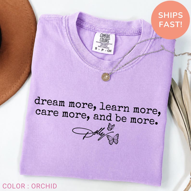 May include: A light purple short-sleeved t-shirt featuring the black printed text "dream more, learn more, care more, and be more." below which is the name "Dolly" in a script font accompanied by two small black butterfly illustrations.