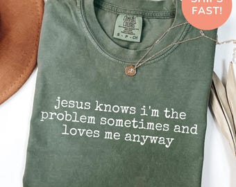 Könnte beinhalten: Ein moosgrünes Kurzarm-T-Shirt mit Rundhalsausschnitt. Das T-Shirt hat einen weißen Textaufdruck auf der Vorderseite: jesus knows i'm the problem sometimes and loves me anyway. Eine dünne goldfarbene Halskette liegt auf dem T-Shirt.