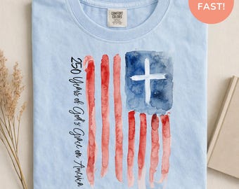 Könnte beinhalten: Ein hellblaues T-Shirt mit Rundhalsausschnitt und einer Aquarell-Grafik einer amerikanischen Flagge mit einem weißen Kreuz im blauen Feld. Der Text 250 Years of God's Grace on America ist vertikal in schwarzer Schrift links neben der Flagge gedruckt.