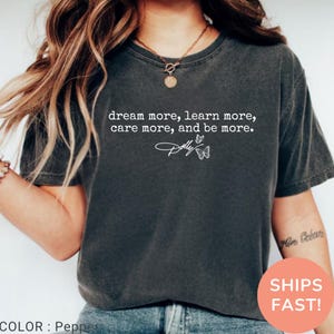 May include: A charcoal grey short-sleeve t-shirt featuring white text that reads dream more, learn more, care more, and be more. Below the text is the word Dolly in a script font with two small butterfly illustrations on the right side.