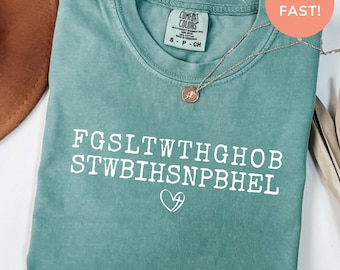 Könnte beinhalten: Meergrünes T-Shirt mit weißem Text und einem Herz mit Kreuz. Das Shirt hat einen Rundhalsausschnitt und kurze Ärmel. Eine silberne Halskette mit Anhänger liegt über dem Shirt. Das Shirt ist auf einer weißen Oberfläche gefaltet.
