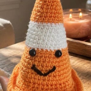 May include: A small, cone-shaped crochet object featuring a bright orange base and top section separated by a horizontal white band. The cone has two round black plastic eyes and a stitched black yarn mouth, creating a simple, friendly facial expression.