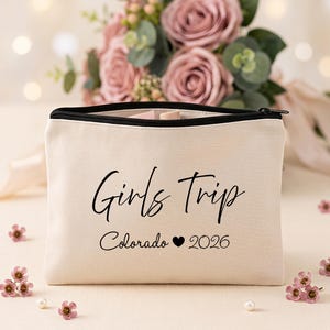 May include: A beige canvas zippered pouch featuring the black printed text Girls Trip, Colorado, a heart symbol, and the year 2026. The rectangular bag has a black zipper closure along the top edge and a structured, flat-bottomed design.