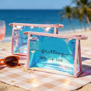 May include: Two rectangular iridescent pouches with pink trim. The front pouch features the black text "Brittany" in script above "Girl's Trip 2026" in a sans-serif font. The pouches have a semi-transparent, holographic finish that reflects light.