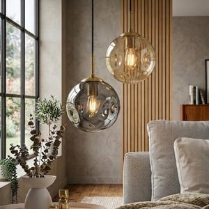 May include: Two spherical glass pendant lights with textured surfaces and brass-toned fittings. One is clear, the other smoky grey. Each has a visible lightbulb, suspended from a black cord. The lights hang in a living room setting.