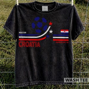 Puede incluir: Una camiseta negra de manga corta con un gr&aacute;fico que muestra un bal&oacute;n de f&uacute;tbol azul, rayas rojas y blancas, y el texto "SOCCER CROATIA". El dise&ntilde;o incluye una peque&ntilde;a bandera de Croacia, dos estrellas y el texto "WORLD CUP '26".