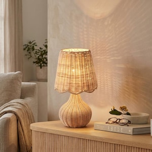 May include: A woven rattan table lamp with a scalloped lampshade. The lamp sits on a wooden nightstand with a book, glasses, and a mug. The lamp casts a warm glow, creating a cozy atmosphere. The lamp is a decorative home accent.