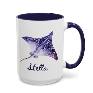 May include: A white ceramic mug with a dark blue interior and handle. The exterior features a purple and white spotted stingray illustration positioned above the name Stella written in a dark blue cursive script font on the front surface.
