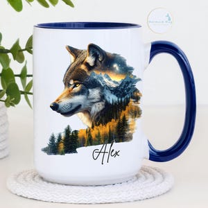 May include: A white ceramic mug with a dark blue interior and handle. The exterior features a graphic of a wolf head integrated with a mountain landscape, pine trees, and a starry sky. The name Alex is printed in black script below the graphic.