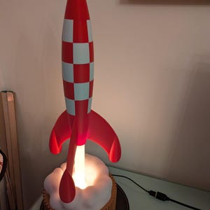 Könnte beinhalten: Eine rot-weiß karierte Raketenschiff-Lampe mit roten Flossen. Die Rakete steht auf einer weißen, wolkenartigen Basis, aus der ein warmes Licht austritt. Die Lampe ist an eine Steckdose angeschlossen.